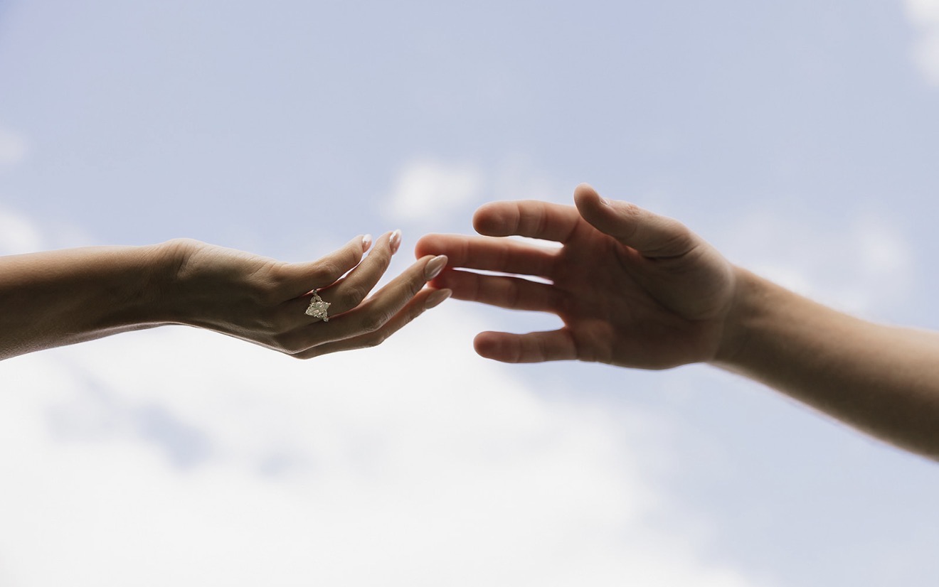 Two hands reaching toward each other against an open sky, one wearing a custom engagement ring